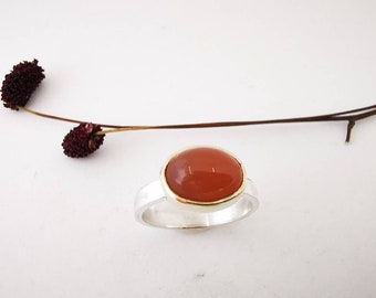 May include: A silver ring with a bezel setting featuring a smooth, oval-shaped carnelian gemstone. The carnelian is a warm orange-red color.