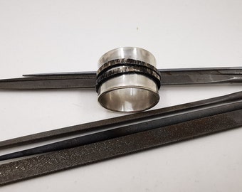 May include: A silver ring with two black bands around it, sitting on a white surface with several metal files.