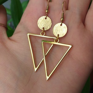 Può includere: Orecchini geometrici color oro. Ogni orecchino presenta un perno circolare strutturato e un ciondolo triangolare. Gli orecchini sono sospesi da ganci per orecchie color oro. Gli orecchini sono esposti su uno sfondo sfocato.