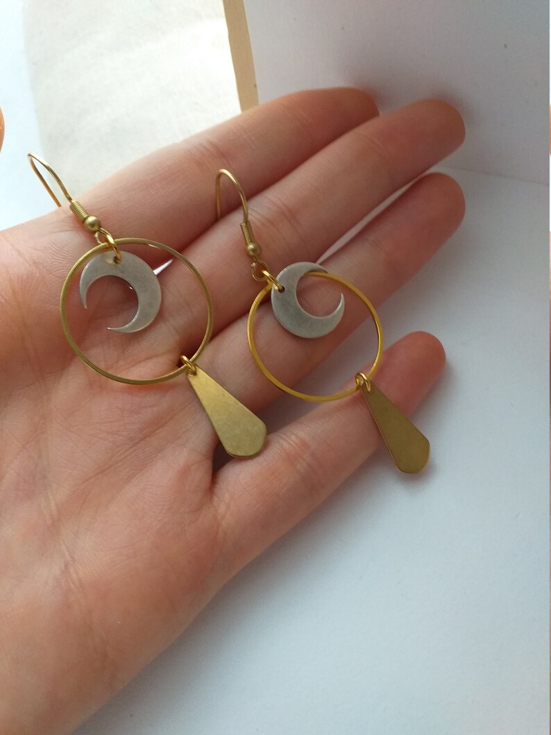 Op de afbeelding: Een paar goudkleurige oorbellen met een maanmotief. Elke oorbel heeft een gouden ring, een zilveren halve maan en een druppelvormige hanger. De oorbellen worden gepresenteerd tegen een lichte achtergrond.