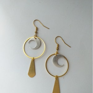Op de afbeelding: Een paar goudkleurige oorbellen met een hemels thema. Elke oorbel heeft een zilveren halve maan in een gouden ring, met een gouden druppelhanger eronder. De oorbellen staan tegen een witte achtergrond.