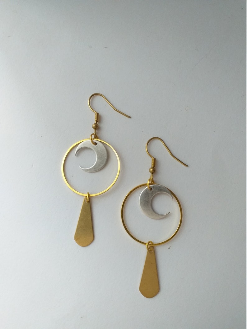 Op de afbeelding: Een paar goudkleurige oorbellen met een hemels thema. Elke oorbel heeft een zilveren halve maan in een gouden ring, met een gouden druppelhanger eronder. De oorbellen staan tegen een witte achtergrond.