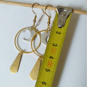 Op de afbeelding: Goud- en zilverkleurige oorbellen met een halve maan en druppelvormige hangers. De oorbellen worden gemeten met een gele meetlint. De oorbellen zijn ongeveer 5 cm lang.