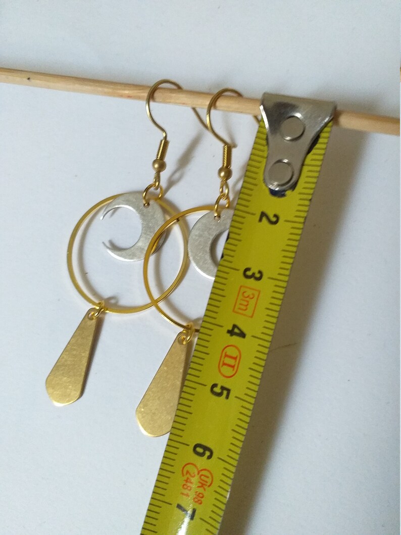 Op de afbeelding: Goud- en zilverkleurige oorbellen met een halve maan en druppelvormige hangers. De oorbellen worden gemeten met een gele meetlint. De oorbellen zijn ongeveer 5 cm lang.