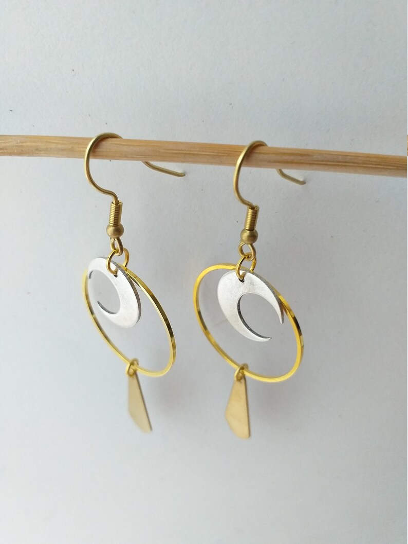Op de afbeelding: Een paar goudkleurige oorbellen met een hemels thema. Elke oorbel heeft een gouden ring, een zilveren halve maan en een kleine gouden driehoekige hanger. De oorbellen zijn opgehangen aan goudkleurige oorhaken.