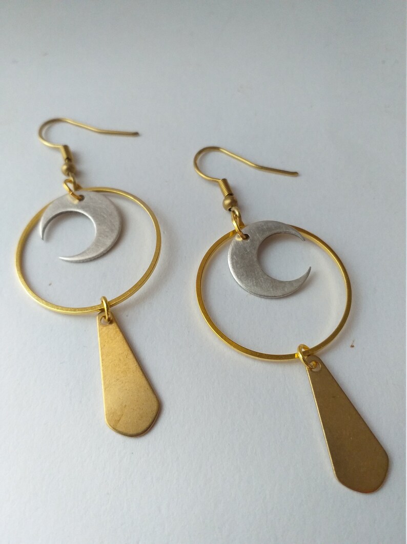Op de afbeelding: Een paar goudkleurige oorbellen met een halve maan ontwerp. Elke oorbel heeft een goudkleurig cirkelvormig frame, een zilveren halve maan en een goudkleurige druppelvormige hanger. De oorbellen worden gepresenteerd tegen een witte achtergrond.