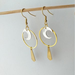 Op de afbeelding: Een paar goudkleurige oorbellen met een zilveren halve maan in een gouden cirkel. Onder de cirkel hangt een gouden druppelvorm. De oorbellen worden gepresenteerd op een houten stok tegen een witte achtergrond.