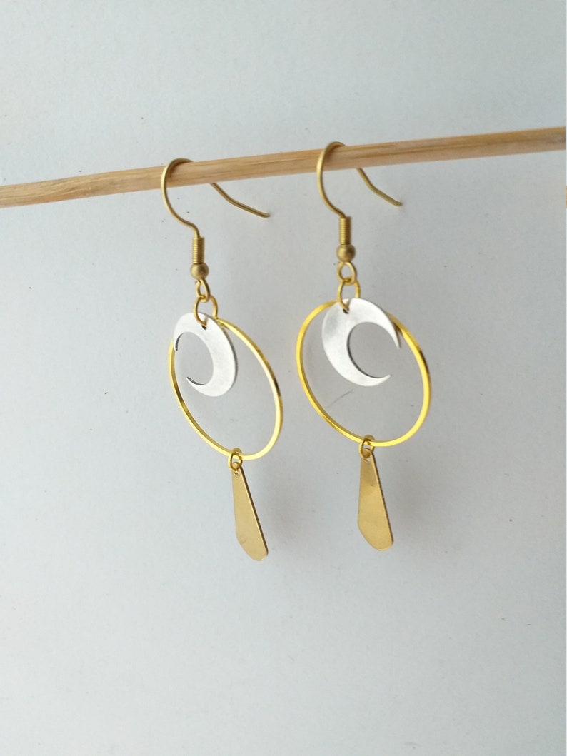 Op de afbeelding: Een paar goudkleurige oorbellen met een zilveren halve maan in een gouden cirkel. Onder de cirkel hangt een gouden druppelvorm. De oorbellen worden gepresenteerd op een houten stok tegen een witte achtergrond.