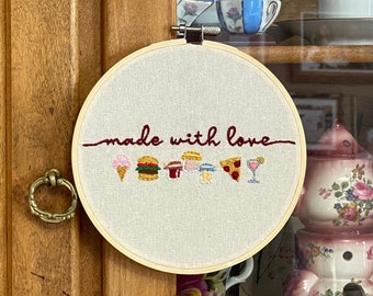 May include: A white embroidery hoop with the text "made with love" in red cursive. The hoop features a line of embroidered food icons including a burger, ice cream cone, pizza slice, and a cocktail glass.