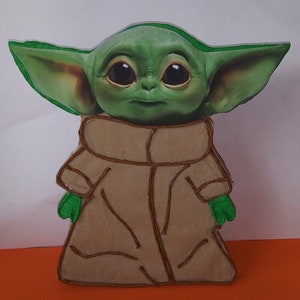 May include: A green and brown felt cutout of a character from the Star Wars franchise, known as "The Child" or "Baby Yoda". The character is wearing a brown robe and has large eyes.