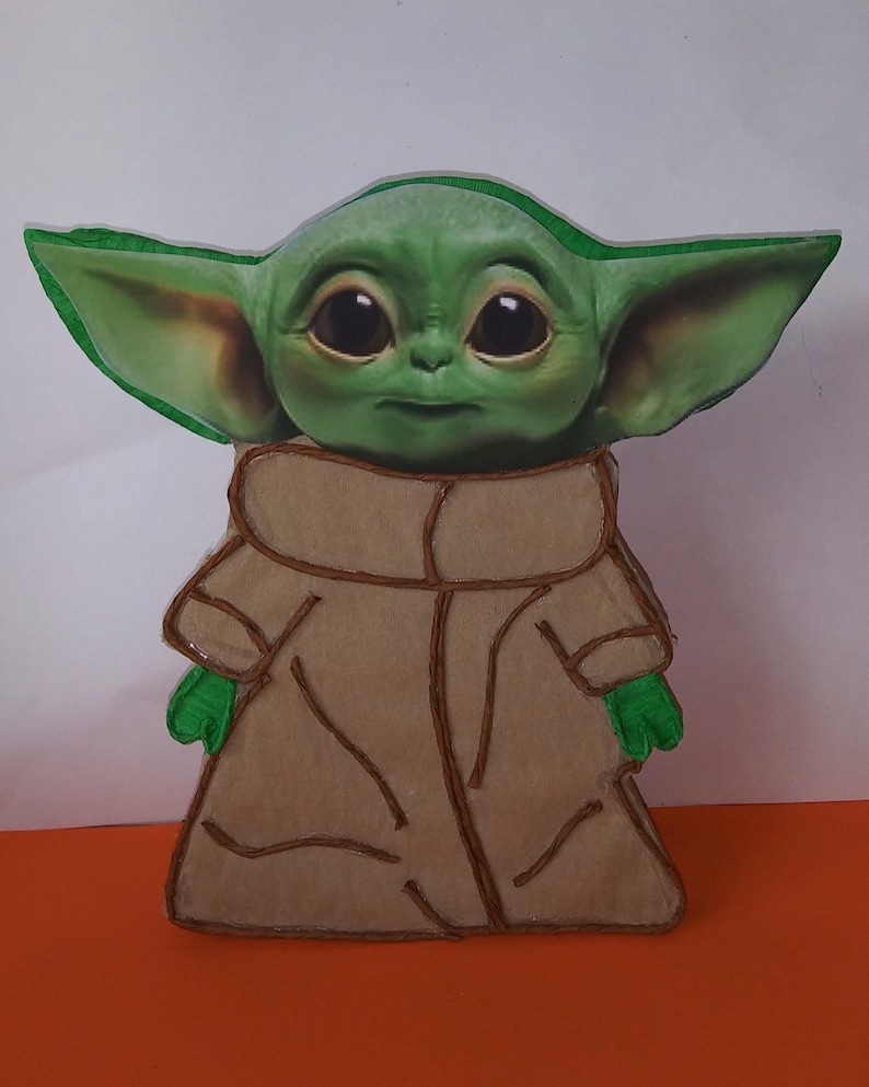 May include: A green and brown felt cutout of a character from the Star Wars franchise, known as "The Child" or "Baby Yoda". The character is wearing a brown robe and has large eyes.