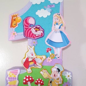 May include: A colorful cardboard pinata shaped like the number one, decorated with characters from Alice in Wonderland, including Alice, the Cheshire Cat, the Mad Hatter, and the White Rabbit. The pinata has a blue background with white clouds and pink and purple accents. The text "Drink Me" is visible on the pinata.