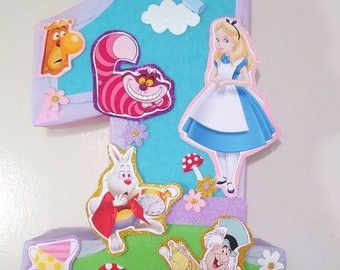 Op de afbeelding: Een kleurrijke kartonnen piñata in de vorm van het nummer één, versierd met personages uit Alice in Wonderland, waaronder Alice, de Cheshire Cat, de Mad Hatter en de Witte Konijn. De piñata heeft een blauwe achtergrond met witte wolken en roze en paarse accenten. De tekst "Drink Me" is zichtbaar op de piñata.