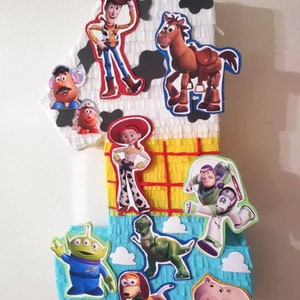 May include: A colorful, multi-layered piñata shaped like the number one, decorated with characters from the Toy Story movie franchise. The piñata features Woody, Buzz Lightyear, Jessie, Bullseye, Mr. Potato Head, Rex, Slinky Dog, and Hamm.