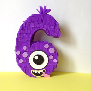 May include: A purple number six piñata in a monster design. The piñata is covered in fringed paper and features a large eye, small purple circles, and a smiling mouth with a pink tongue. A fun party decoration.