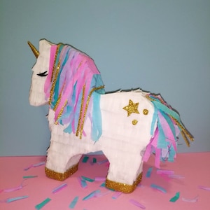 May include: A white unicorn pinata with a gold star and gold glitter accents. The pinata has pink, blue, and white tissue paper streamers.