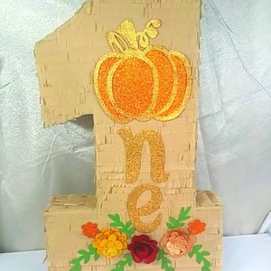 May include: A brown paper mache number one piñata with a gold glitter one and a pumpkin with orange glitter on it. The piñata is decorated with green leaves and red, orange, and yellow flowers.