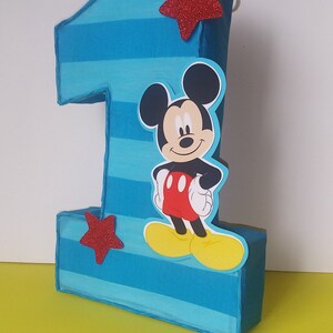 Puede incluir: Pi&ntilde;ata de cart&oacute;n n&uacute;mero uno con rayas azules y blancas, una estrella roja y un personaje de Mickey Mouse en la parte delantera.