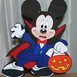 May include: A Halloween-themed cutout of Mickey Mouse dressed as a vampire, wearing a blue suit with a red bow tie and cape. He holds a jack-o'-lantern bucket. The character has white gloves and a smiling expression.