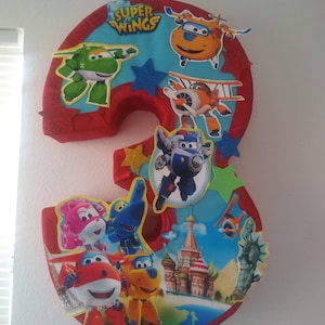 May include: A red number three piñata decorated with characters from the Super Wings cartoon. The characters include Jett, Dizzy, Jerome, Paul, and Mira. The piñata also features a red star, a blue star, and a green star.