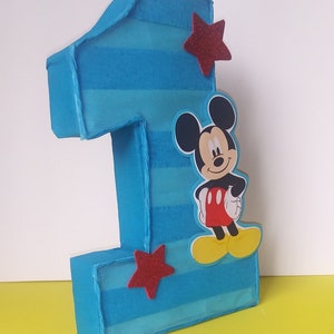Puede incluir: Un n&uacute;mero uno de cart&oacute;n a rayas azules y blancas con una estrella roja y un personaje de Mickey Mouse.