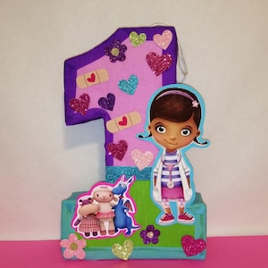 May include: A pink and purple number one shaped pinata with a cartoon image of Doc McStuffins, a blue dragon, and a white sheep. The pinata is decorated with glitter, hearts, and band-aids.