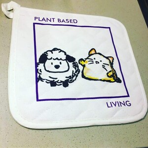 以下が含まれることがあります： 紫色の縁取りと「PLANT BASED」と「LIVING」の文字が入った、白いキルティング鍋つかみ。デザインは漫画の羊と猫が特徴です。鍋つかみには吊り下げ用のループが付いています。