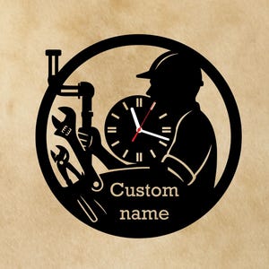 Peut inclure: Horloge murale en métal noir représentant une silhouette de plombier tenant des outils, un tuyau et un cadran d'horloge. L'horloge porte les mots "Custom name" en dessous du cadran. L'horloge est dans un cercle noir.