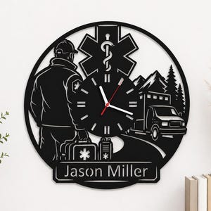 May include: Black metal wall clock featuring an emergency medical technician with an ambulance, mountains, and a medical symbol. The clock hands are red. The name "Jason Miller" is displayed at the bottom.
