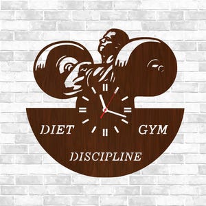 May include: A brown wooden wall clock with a weightlifter design. The clock features a barbell and the words "DIET", "GYM", and "DISCIPLINE". The clock hands are white with a red second hand. The background is a white brick wall.