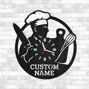 May include: Black metal wall clock featuring a chef silhouette holding a knife and whisk. The clock face is integrated into the chef's body, with the text "CUSTOM NAME" at the bottom. The clock is set against a white brick wall.
