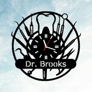 Op de afbeelding: Zwarte metalen wandklok met een tandheelkundig thema, met tandheelkundige instrumenten en een tand-silhouet. De wijzerplaat is omgeven door diverse tandheelkundige instrumenten. De naam "Dr. Brooks" staat op een banner eronder. De wijzers zijn zwart met een rode secondewijzer.