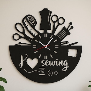 Könnte beinhalten: Schwarze Wanduhr mit Näh-Motiv. Die Uhr zeigt ein Herz, die Worte "I love sewing" und Bilder von Nähwerkzeugen wie Schere, Maßband und Schneiderpuppe. Die Uhrzeiger sind weiß mit einem roten Sekundenzeiger.