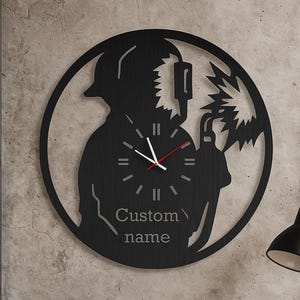Peut inclure: Une horloge murale en métal noir représentant la silhouette d'un soudeur dans un cadre circulaire. Les aiguilles de l'horloge sont blanches et rouges, et le cadran porte l'inscription "Custom name". L'horloge est fixée à un mur gris texturé.