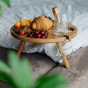 May include: A wooden picnic table with a round top and three legs. The table holds a croissant, apricots, strawberries, and cherries. A bottle of champagne and two glasses are also on the table.