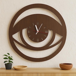 May include: A brown wooden wall clock in the shape of an eye. The clock face is circular with a smaller clock face inside. The hands are white with a red second hand. The clock is mounted on a white wall above a wooden cabinet.