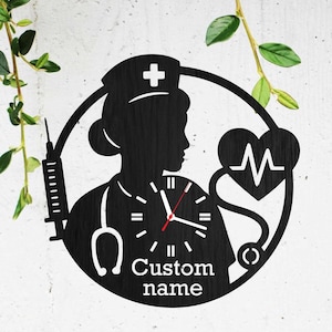 May include: Black metal wall clock featuring a nurse silhouette within a circular frame. The design includes a syringe, stethoscope, and a heartbeat symbol. The clock face displays the text "Custom name".