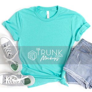 May include: A turquoise t-shirt with rolled sleeves, paired with light-wash denim shorts and gray canvas sneakers. The t-shirt is knotted at the waist. The text "THE TRUNK STUDIO Mockups" is visible.
