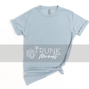 Op de afbeelding: Een lichtblauw t-shirt met opgerolde mouwen, geknoopt aan de onderkant. Het shirt heeft een ronde hals en een label bij de kraag. De tekst "THE TRUNK STUDIO Mockups" staat over het midden van het shirt.
