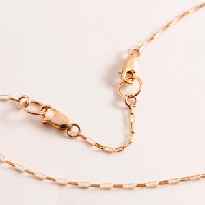May include: A delicate gold-colored necklace with a lobster clasp and an adjustable chain. The chain is composed of small, rectangular links. The necklace is displayed on a white surface, highlighting its fine details.
