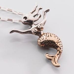 May include: A silver mermaid pendant with a gold tail, hanging from a fine chain. The pendant features a mermaid with flowing hair and outstretched arms. The design is whimsical and elegant, suitable for everyday wear.