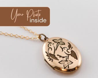 May include: An oval gold-toned locket pendant on a delicate gold-toned chain. The front of the locket features an engraved design of two birds perched among flowers. A brown rectangular graphic overlay contains the white text Your Photo inside.