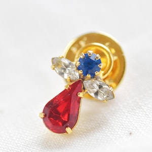 Könnte beinhalten: Eine goldfarbene Brosche mit einem roten, blauen und weißen Kristalldesign. Die Brosche hat einen tropfenförmigen roten Kristall, einen runden blauen Kristall und zwei weiße Kristalle in Marquise-Form.