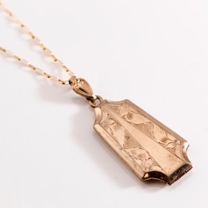 May include: A gold-colored pendant necklace with an ornate, rectangular pendant. The pendant features engraved floral designs and a central, vertical panel. The chain is delicate and gold-toned.