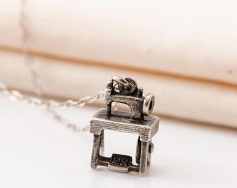 May include: A silver-colored sewing machine pendant necklace. The pendant is a detailed miniature of a vintage sewing machine, mounted on a small table. The necklace chain is also silver. The background is a soft, neutral color.