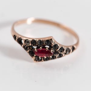 Pode incluir: Um anel de ouro rosa com uma pedra preciosa vermelha oval no centro, ladeada por pedras pretas. O anel tem um design único e decorativo, sobre um fundo branco. Uma joia.