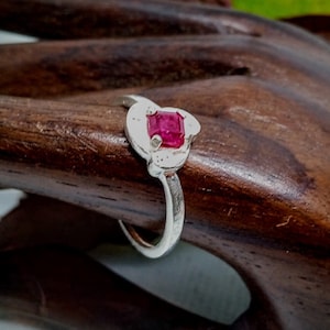 Könnte beinhalten: Ein silberner Ring mit einem quadratischen, leuchtend roten Edelstein in einer dekorativen Fassung. Der Ring wird auf einer dunklen Holzoberfläche präsentiert, um sein Design und seine Farbe hervorzuheben.