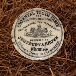 Puede incluir: Una tapa de cerámica blanca para un tarro de pasta dental oriental. La tapa tiene texto negro que dice "Oriental Tooth Paste for Cleansing Beautifying and Preserving the Teeth & Gums. Prepared by Jewsbury & Brown Chemists 113 Market Street Manchester".