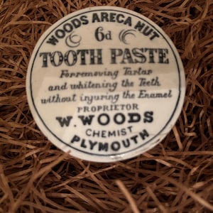 Op de afbeelding: Een wit keramiek deksel voor een tandpasta pot. Het deksel heeft zwarte tekst die "Woods Areca Nut 6d Tooth Paste Forremoving Tartar and whitening the Teeth without injuring the Enamel Proprietor W. Woods Chemist Plymouth" luidt.