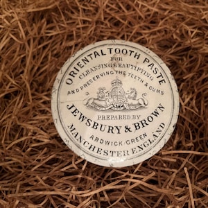 Może przedstawiać: Okrągła, zabytkowa puszka "Oriental Tooth Paste" firmy Jewsbury & Brown. Biała puszka ma czarny tekst i herb z napisem "Cleansing, Beautifying, and Preserving the Teeth & Gums". Puszka leży na brązowej słomie.